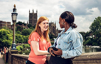 2 students stood smiling and facing each other on 91 bridge, with 91 Cathedral in the background